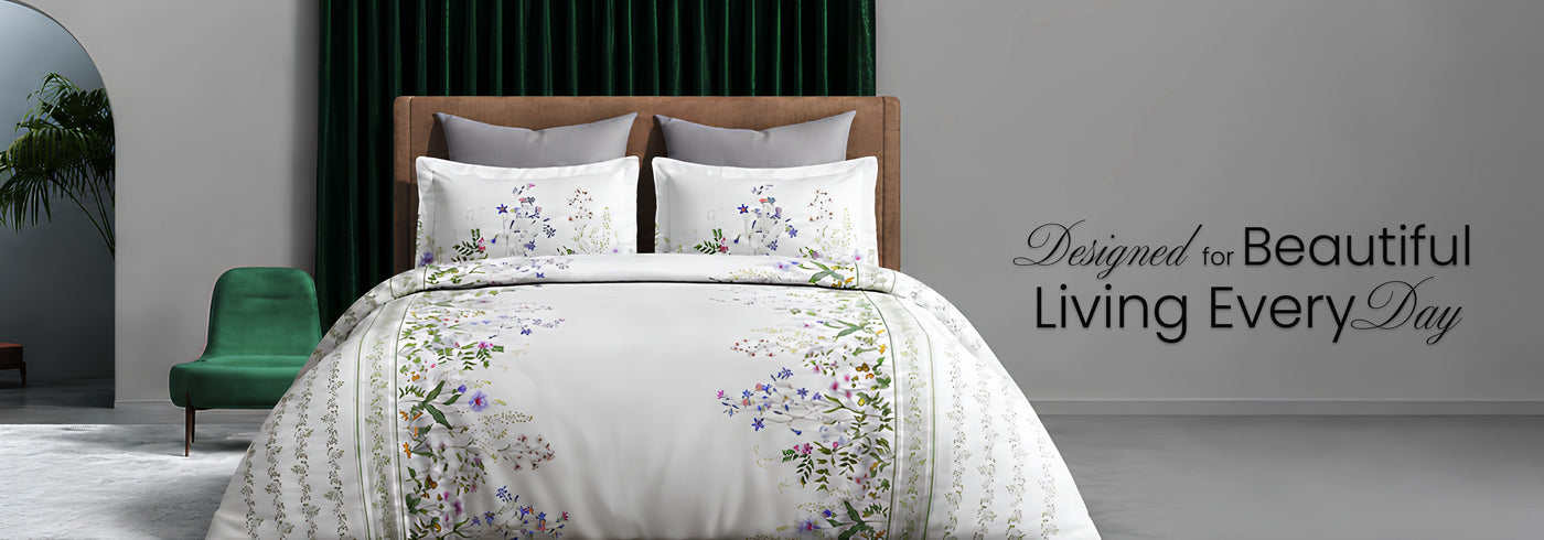 A styled bedroom scene showing a bed dressed in white bedding with a floral border print, set against a dark green curtain and a gray wall. A green chair is visible on the left, and the text 'Designed for Beautiful Living Every Day' appears on the right.